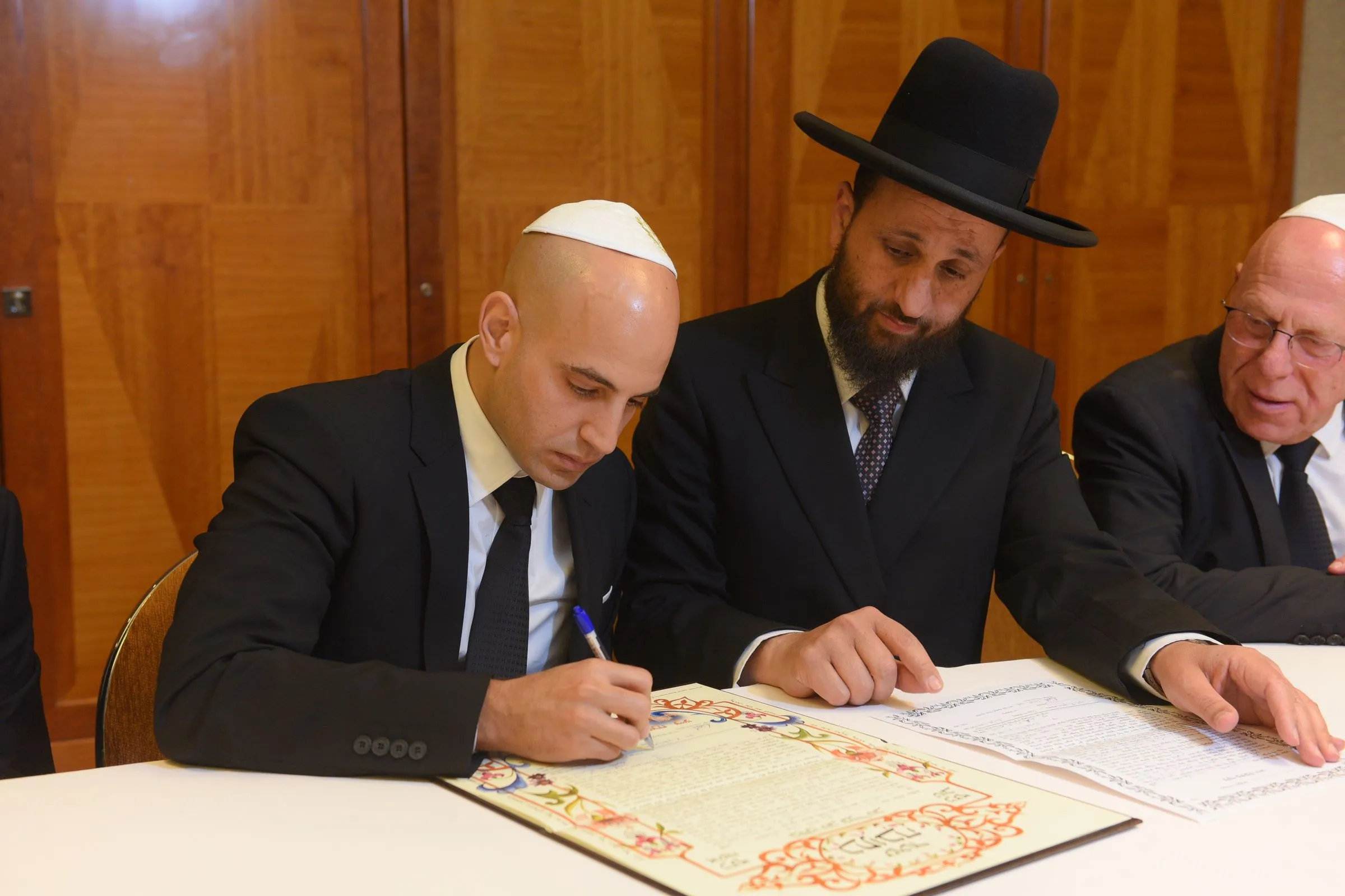 A man in a white kippah signs an illuminated Hebrew document at a table while a man in a black hat and an older man in a kippah look on