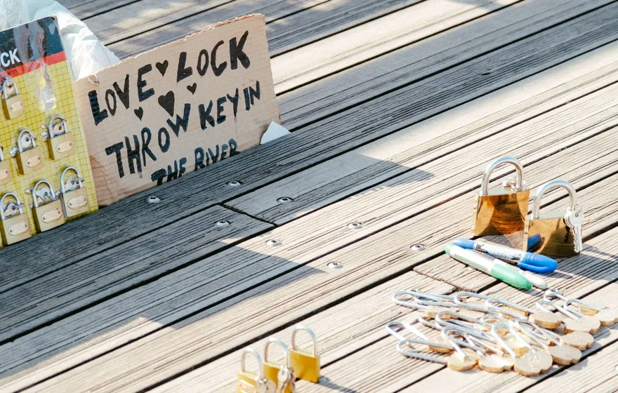 A cardboard sign reading Love Lock, Throw Key in the River propped on a wooden boardwalk beside a display of padlocks for sale, marker pens and a few locks on the boards