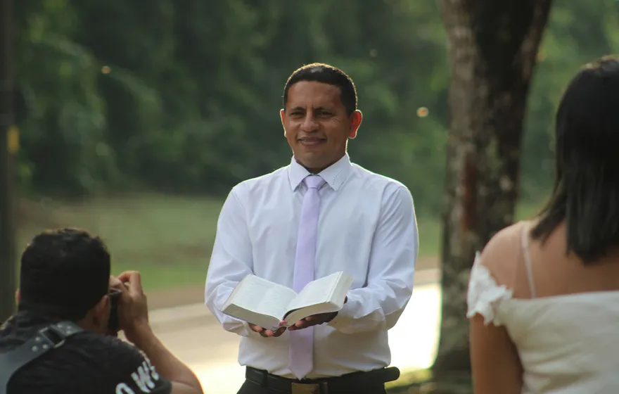 An officiant in a white shirt and tie stands outdoors holding an open book, guests seated in the foreground