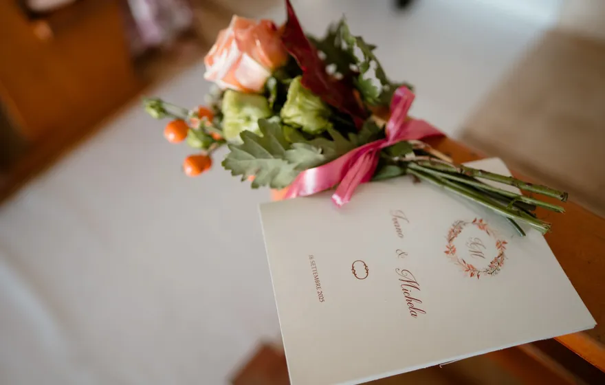 A folded cream ceremony program printed with the couple's names and a pink floral monogram, lying on a wooden seat under a small bouquet of roses and greenery tied with pink ribbon