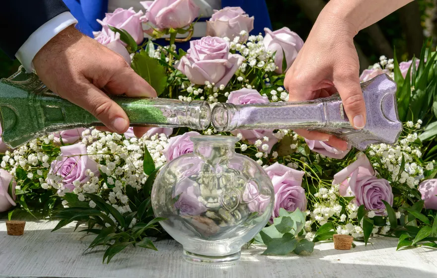 Two hands pour lavender sand from long glass vials into a round glass vase set among purple roses and baby's breath