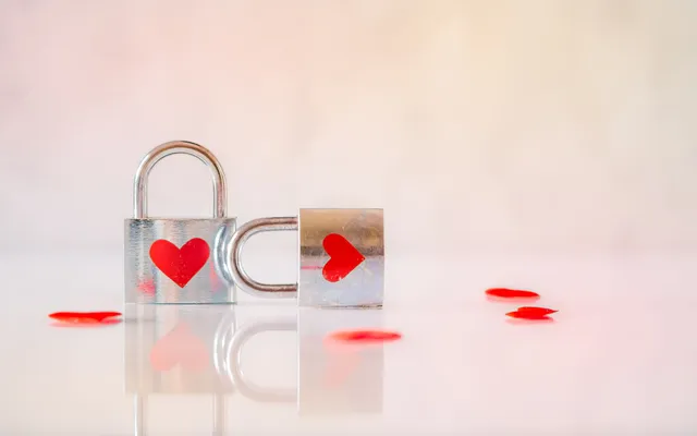 Two silver padlocks with red hearts on a white tabletop, the shackle of one hooked through the other, red petals scattered around them