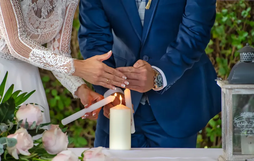 A bride and groom each hold a thin lit taper to a white pillar candle on a table decorated with pink roses